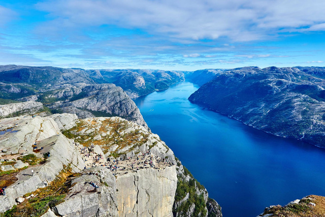 A flat-topped granite cliff dotted with small figures overlooks a deep blue fjord winding between rugged mountains under a partly cloudy sky.