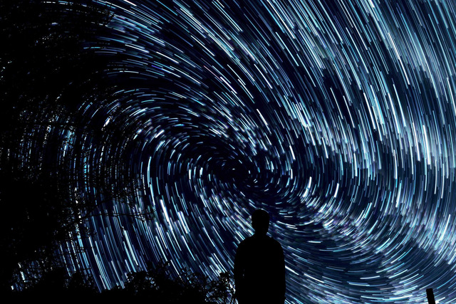 A long-exposure night photograph showing concentric arcs of star trails curving across a dark sky, with a silhouetted person in the lower right and dark tree foliage framing the left side.