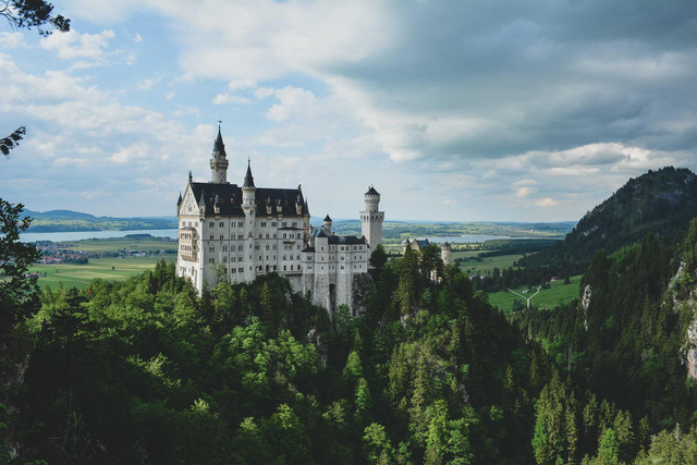A large white stone castle with multiple towers and a tall central spire sits atop a forested ridge, with a valley of green fields, scattered settlements, and distant mountains visible behind it.