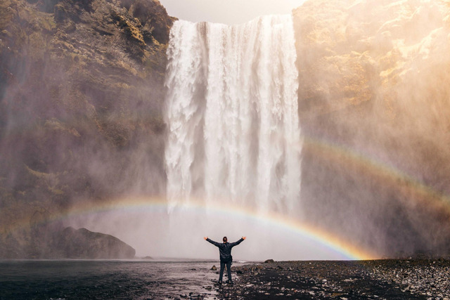 A figure with arms outstretched faces a tall waterfall from a pebbled shoreline, with a full rainbow arcing across the mist in warm golden light.